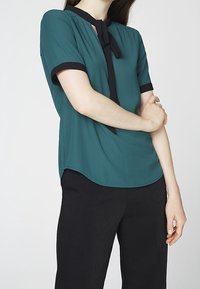 Blusa teal con maniche corte, rifinitura nera e dettaglio del collo legato; abbinata a pantaloni neri. Tessuto liscio con una leggera lucentezza.