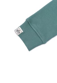 Teal sweatshirtärm med ribbad mudd, slät textur och en märkesetikett med en geometrisk design på sidan.