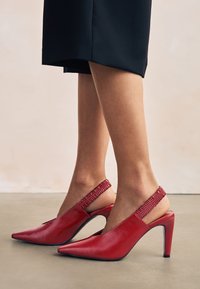 Escarpins slingback en cuir rouge avec un bout pointu, un talon étroit et une sangle élastique tressée à la cheville. Surface lisse, design élégant.