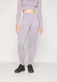 Svijetloljubičasti dugih rukava crop top u kombinaciji s uskladenim visokim tajicama. Materijal je gladak i rastezljiv. Završeno s crnim sportskim cipelama.