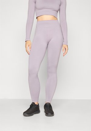 Svijetloljubičasti dugih rukava crop top u kombinaciji s uskladenim visokim tajicama. Materijal je gladak i rastezljiv. Završeno s crnim sportskim cipelama.