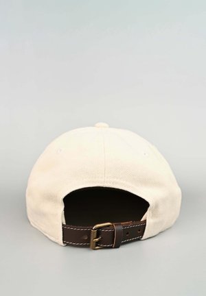 Beżowa czapka z daszkiem widziana od tyłu z regulowanym ciemnobrązowym skórzanym paskiem i metalową sprzączką na jednolitym jasnym tle.