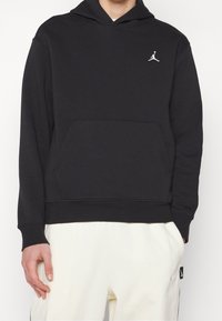 Schwarzer Kapuzenpullover aus Baumwollmischgewebe, mit Kängurutasche und weißem Jumpman-Logo auf der Brust. Glatte Textur, lockere Passform.