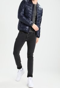 Doudoune marine avec fermeture éclair sur le devant, pull gris, jeans noirs slim et baskets blanches. La doudoune présente une texture matelassée.