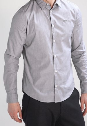 Man draagt een lichtgrijs, overhemd met lange mouwen en knoopsluiting, en zwarte broek, staand tegen een effen lichte achtergrond.