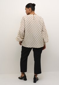 Beige blus med svarta prickar, långa ballongärmar och en nyckelhålsdetalj på baksidan, ihop med svarta smala byxor och klackskor.