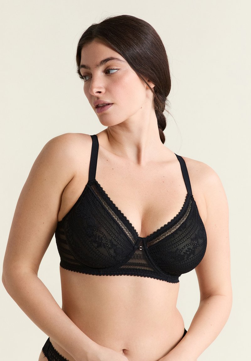 Mujer con el cabello oscuro trenzado, usando un sujetador negro de encaje con aro, mirando hacia su izquierda sobre un fondo claro liso.