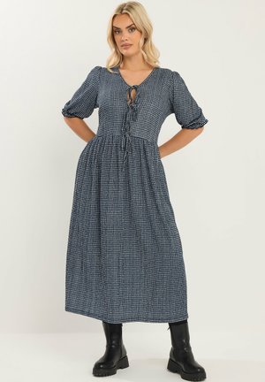Femme portant une robe midi à carreaux bleu marine et blanc avec des manches bouffantes et des bottines noires, debout les mains sur les hanches.