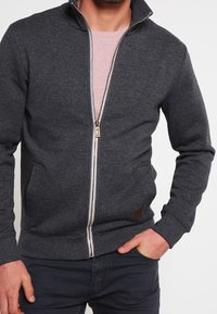 Sudadera con cremallera de color gris oscuro, con cuello alto, cremallera dorada, dos bolsillos frontales, puños de canalé y pequeño parche de cuero marrón con el logotipo.