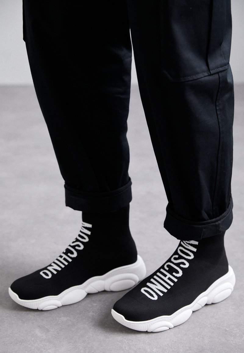 Čierne tenisky v štýle ponožiek s bielymi podrážkami. Na bokoch sa nachádza biele brandovanie "MOSCHINO". Nosí sa s srolovanými čiernymi nohavicami.