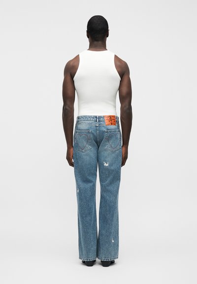 Άνδρας που φοράει άσπρο αμάνικο τοπ και μπλε τζιν με καρδιόσχημες ραφές στις πίσω τσέπες, στέκεται πλάγια με την πλάτη προς τα εμπρός σε απλό φόντο.