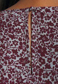 Encolure arrière d'un chemisier à motif floral en bordeaux et gris, dévoilant la peau à travers une ouverture en forme de goutte d'eau fermée par un bouton en tissu.