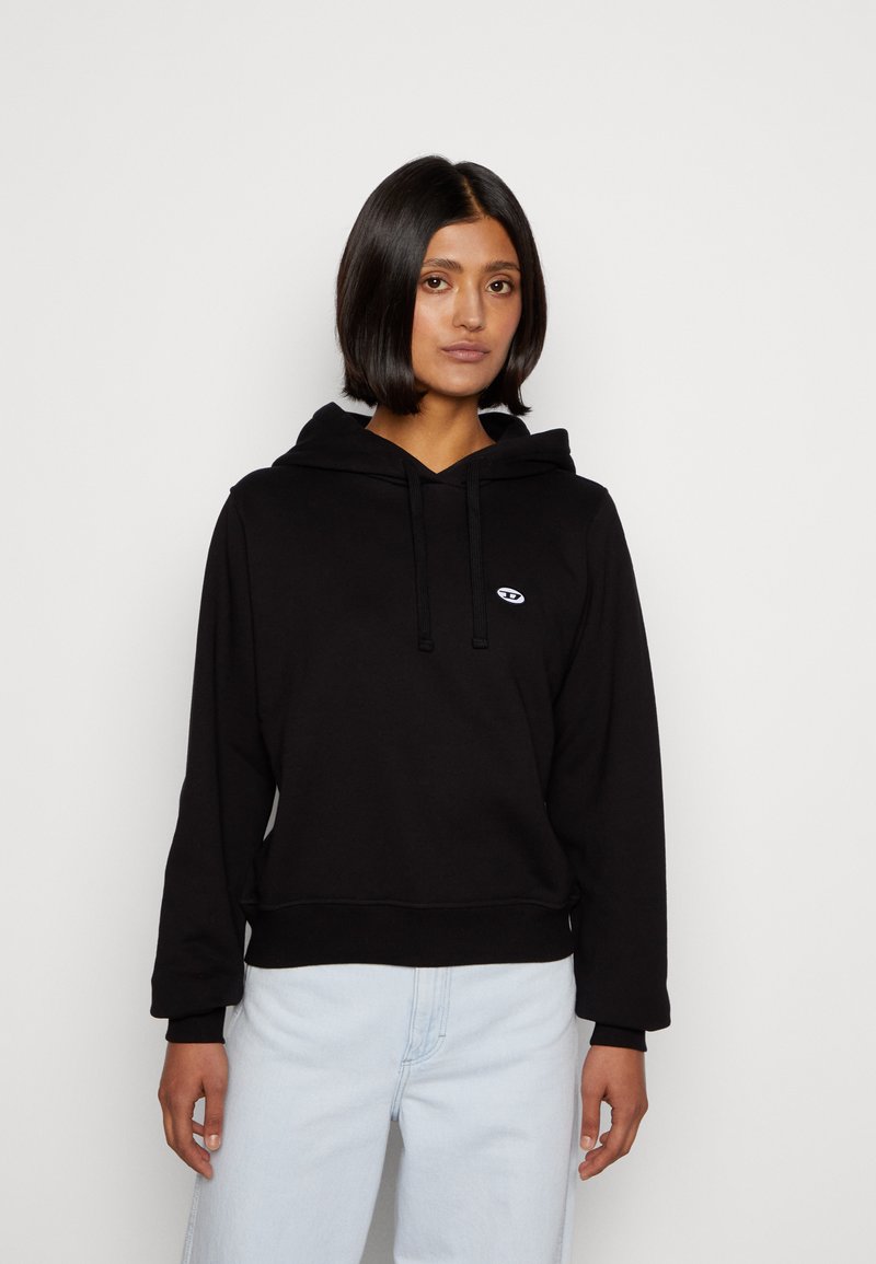 Schwarzer Hoodie aus Baumwolle, ausgestattet mit einem Kordelzug, gerippten Bündchen und einem kleinen Logo auf der Brust. Kombiniert mit hellblauen Jeans.