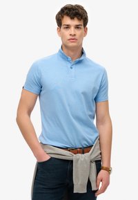 Lichtblauw polo-shirt met een korte opstaande kraag, korte mouwen en een gestructureerde stof; gecombineerd met donkere jeans en een grijze trui die om de taille is geknoopt.