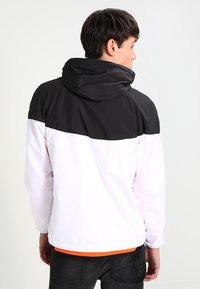 Veste à capuche blanche et noire avec un design coloré, texture lisse, poignets élastiques et un détail orange contrastant à l'ourlet.