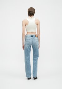 Persoană cu părul scurt, îmbrăcată cu un top scurt alb, blugi decolorati cu talie înaltă de culoare albastră deschisă și pantofi cu toc negri, stând cu spatele la cameră.