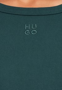 Top ribbed teal cu decolteu rotund și logo-ul brodat "HUGO" într-o nuanță contrastantă. Textură moale și design ajustat.
