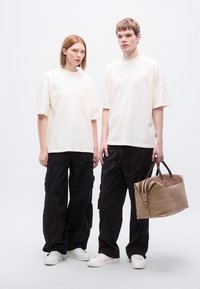 Δύο μοντέλα φορούν χαλαρά, κρεμ T-shirts και μαύρο παντελόνι φορτίου. Ένα κρατάει μια καφέ δερμάτινη τσάντα duffle με σκούρες λεπτομέρειες και φερμουάρ.