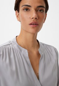 Hellgrau Bluse mit Kragen, plissierten Schulterdetails und einem leichten V-Ausschnitt, aus glattem, glänzendem Stoff gefertigt.