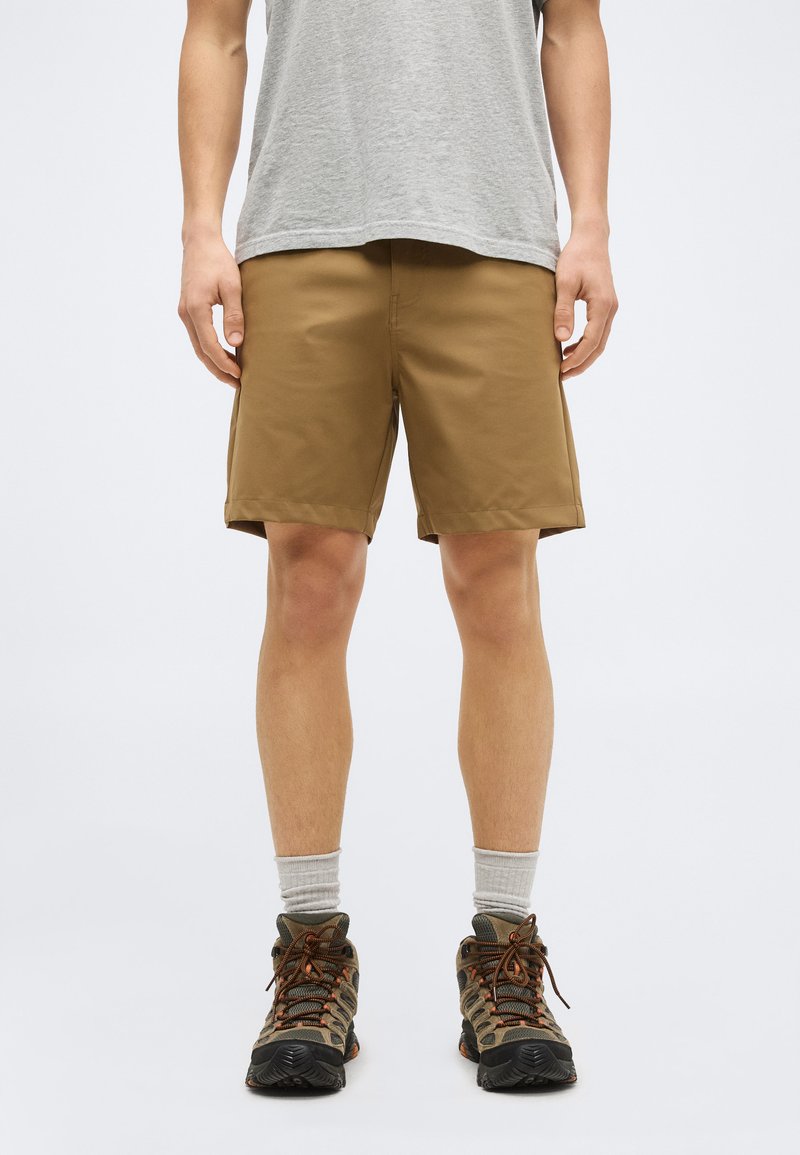 Homme portant un short kaki, un t-shirt gris, des chaussettes grises montantes et des chaussures de randonnée, debout devant un fond blanc uni.