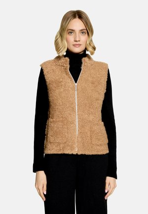 Kvinde iført en beige, blød vest med lynlås over en sort langærmet top og sorte bukser, stående mod en ensfarvet lys baggrund.