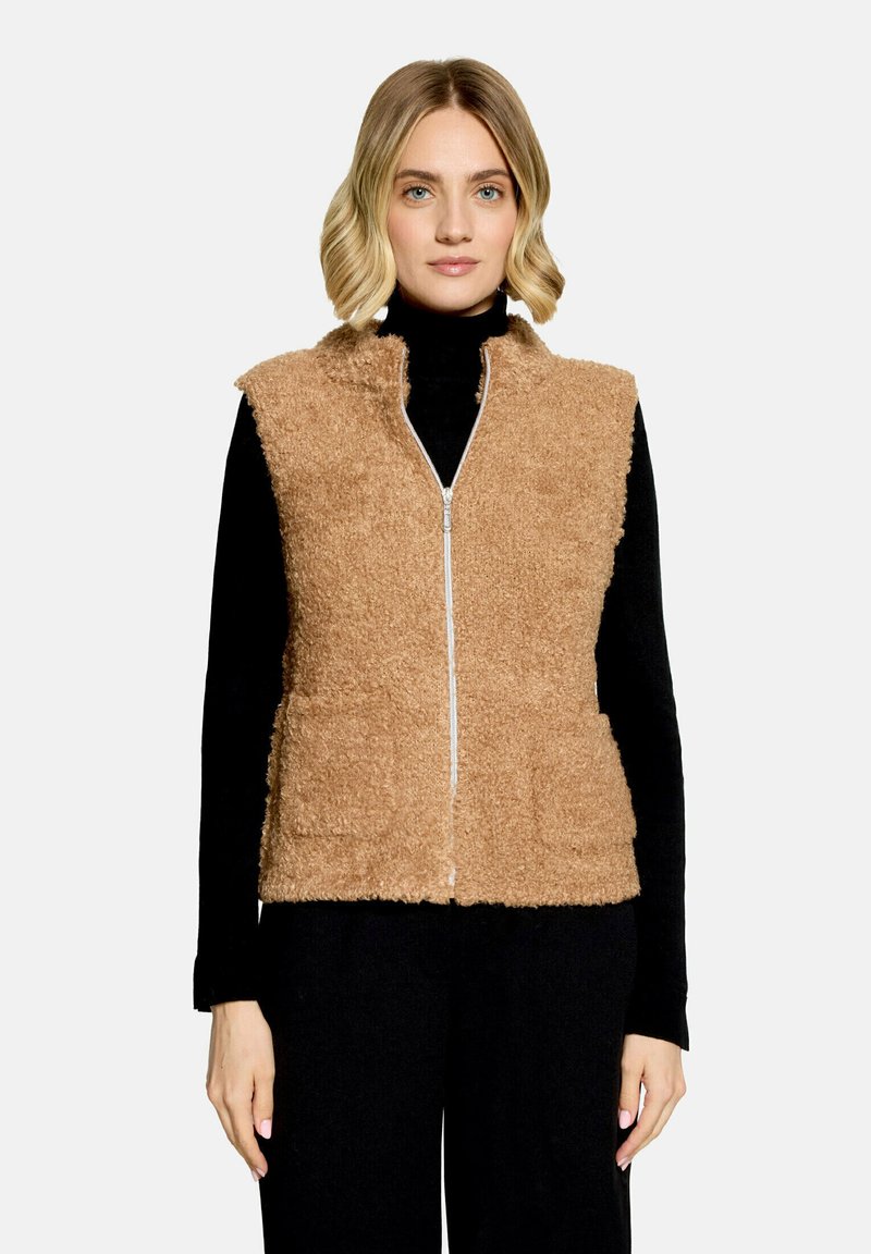 Femme portant un gilet beige duveteux zippé sur un haut noir à manches longues et un pantalon noir, debout devant un fond clair uni.