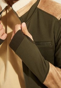 Chaqueta verde oliva con cierre de cremallera, detalles en ante color marrón en los hombros, cuello de forro polar y puños acanalados; cuenta con un pequeño bolsillo con cremallera.