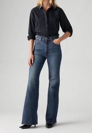 Femme portant une chemise noire boutonnée aux manches retroussées et un jean bleu évasé taille haute, debout avec une main dans la poche.