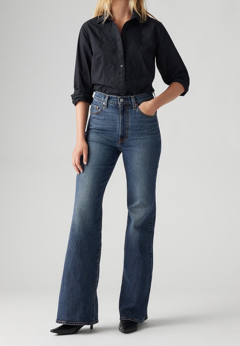 Femme portant une chemise noire boutonnée aux manches retroussées et un jean bleu évasé taille haute, debout avec une main dans la poche.