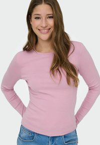 Junge Frau mit langen braunen Haaren, die ein pinkes langärmliges Shirt und blaue Jeans trägt, lächelt mit den Händen auf den Hüften vor einem weißen Hintergrund.