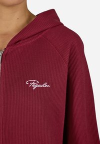Burgundi spalvos su raukšlėmis užtrauktuku užsegama džemperis su gobtuvu, ant krūtinės išsiuvinėtas baltas "Pegador" logotipas. Lygus paviršius ir sportiškas dizainas.