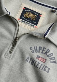 Primer plano de una sudadera gris con cremallera que muestra el texto "Superdry Athletics", una impresión de la bandera estadounidense y una etiqueta cosida en el cuello que dice "Crafted with Passion".
