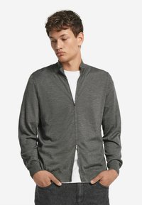 Mörkgrå zip-up cardigan i mjukt tyg, med hög krage, långa ärmar och en smal passform. Bärs över en vit skjorta.