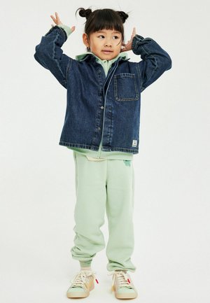 Jeune enfant portant un pantalon vert menthe et des baskets beiges, vêtu d'une veste en jean, posant avec les mains près de la tête sur un fond blanc.