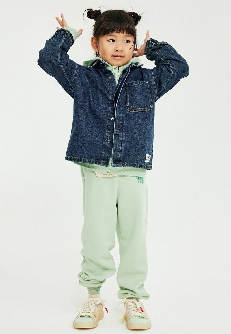 Małe dziecko w miętowo zielonych spodniach i beżowych trampkach, ubrane w kurtkę jeansową, pozujące z rękami przy głowie na białym tle.