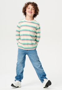 Chemise à manches longues rayée en vert, rouge et bleu sur un fond crème ; elle possède une poche poitrine et un logo. Associée à un jean bleu et des baskets.