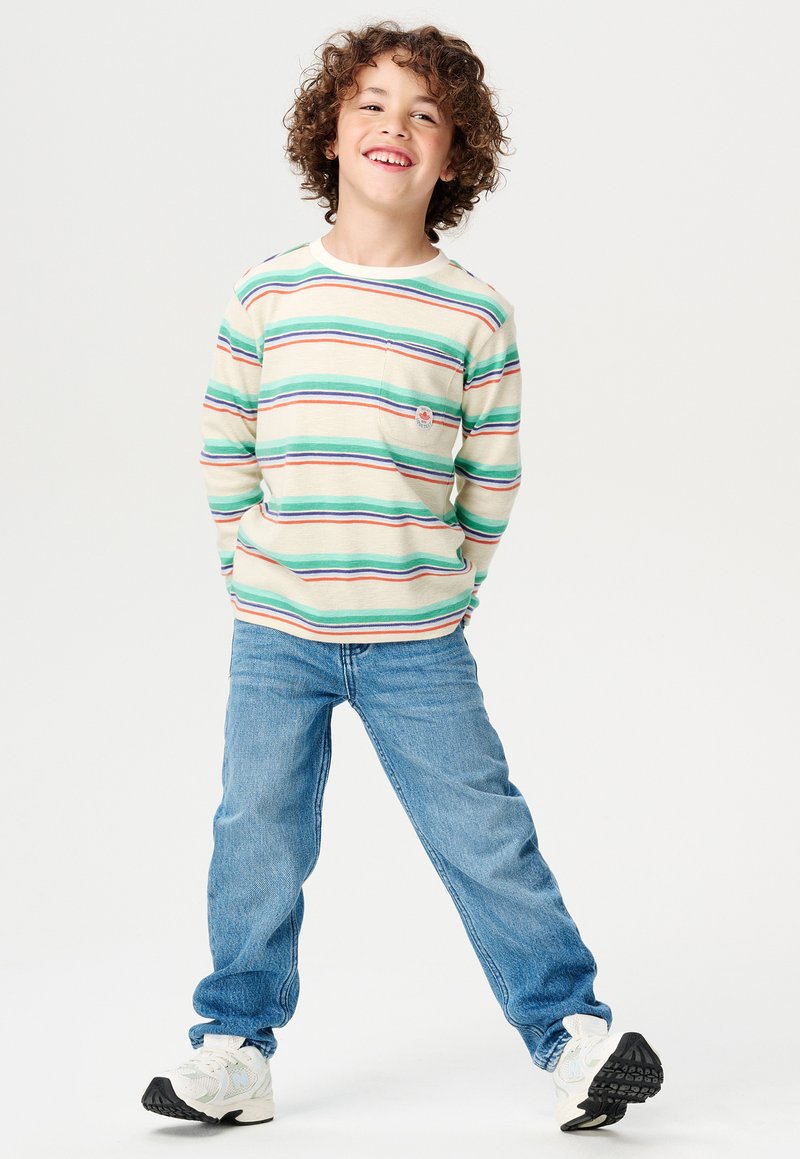 Chemise à manches longues rayée en vert, rouge et bleu sur un fond crème ; elle possède une poche poitrine et un logo. Associée à un jean bleu et des baskets.