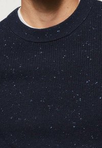 Zbliżenie osoby w ciemnoniebieskim swetrze dzianym z małymi białymi i niebieskimi kropeczkami oraz ściągaczem przy okrągłym dekolcie.