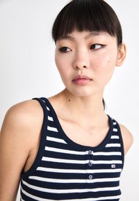 Navy and white striped tank top made of ribbed fabric, featuring a round neckline and three buttons at the front, with a logo detail.