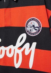 Chemise rayée noire et orange avec texte blanc "Goofy" et un patch circulaire montrant Goofy portant une casquette de baseball avec le texte "Goofin' Around".