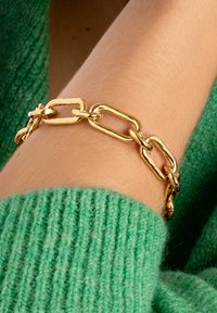 Gouden schakelkettinkje om pols boven een groene gebreide trui, close-up detail van sieraden en textuur.