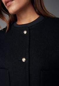 Schwarze Mischwolljacke mit rundem Halsausschnitt, die zwei goldene Knöpfe und eine strukturierte Oberfläche aufweist. Beinhaltet Taschendetails auf der Vorderseite.