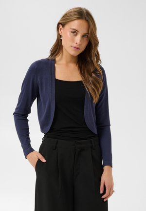 Jeune femme aux longs cheveux châtain clair portant un cardigan court bleu marine sur un haut noir et un pantalon noir, debout avec une main dans la poche.