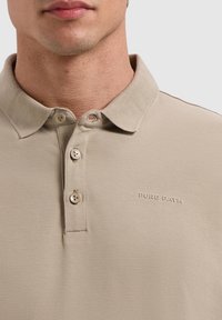 Beige poloshirt met een klassieke kraag, drie knopen en "PURE PATH" geborduurd op de linkerkant van de borst in een subtiele contrasterende kleur.