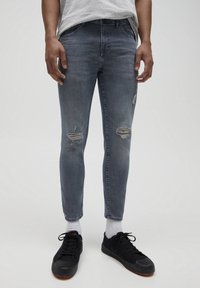 Jeans de denim gris ajustados con desgarros, cintura media y dobladillos sin terminar, combinados con zapatillas negras y calcetines blancos.