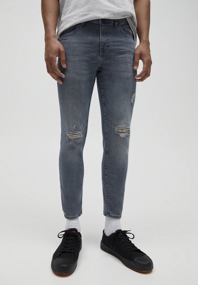 Jeans de denim gris ajustados con desgarros, cintura media y dobladillos sin terminar, combinados con zapatillas negras y calcetines blancos.