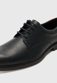 Chaussure en cuir noir avec une texture lisse, design à lacets, et un petit talon. Présente des coutures discrètes et un bout arrondi.