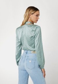 Šviesiai žalia satino bluzelė su baltais taškeliais, ilgomis rankovėmis ir aukštu kaklu. Sukomplektota su šviesiai mėlynomis aukštai talpinamomis džinsomis.