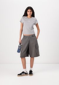 T-shirt cinza cortada com logótipo, calções cargo cinza largos com bolsos, sapatilhas pretas e uma pequena bolsa azul. Fundo branco.