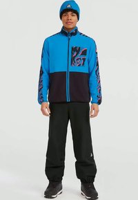 Niebieska kurtka zapinana na zamek z czarnymi akcentami, wzorzystym rękawem i kieszonką na piersi. Połączona z czarnymi spodniami i pasującą niebieską czapką.